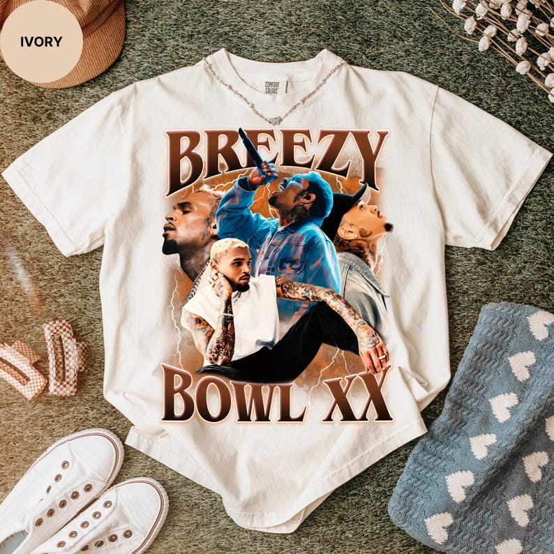 Puede incluir: Camiseta de color marfil con un gr&aacute;fico de un cantante, la palabra "Breezy" arriba y "Bowl XX" abajo. El gr&aacute;fico incluye im&aacute;genes del cantante en varias poses, con acentos de rayos.