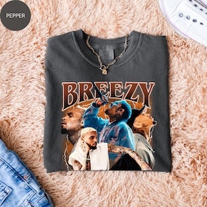 Puede incluir: Camiseta gris oscuro con un gr&aacute;fico de un cantante y la palabra "BREEZY" en marr&oacute;n, con un estilo vintage. La camiseta tiene cuello redondo y mangas cortas. Una cadena de oro est&aacute; sobre la camiseta. La camiseta est&aacute; doblada sobre una superficie color melocot&oacute;n.