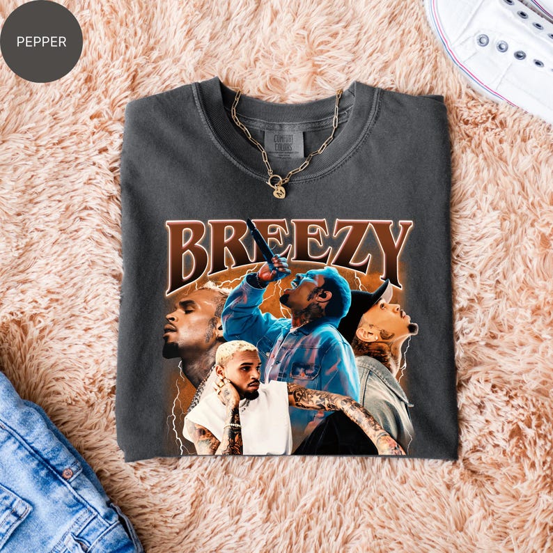 Puede incluir: Camiseta gris oscuro con un gr&aacute;fico de un cantante y la palabra "BREEZY" en marr&oacute;n, con un estilo vintage. La camiseta tiene cuello redondo y mangas cortas. Una cadena de oro est&aacute; sobre la camiseta. La camiseta est&aacute; doblada sobre una superficie color melocot&oacute;n.