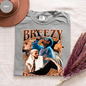 Puede incluir: Una camiseta gris Comfort Colors con un gr&aacute;fico de un cantante y la palabra "BREEZY" en letras marrones. El gr&aacute;fico incluye m&uacute;ltiples im&aacute;genes del cantante con acentos de rayos. La camiseta est&aacute; tendida sobre una superficie blanca.