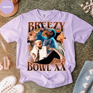 Puede incluir: Una camiseta de color lila claro con un gr&aacute;fico de un cantante, la palabra "BREEZY" arriba y "BOWL XX" abajo. La camiseta tiene un cuello desgastado y mangas cortas. Zapatillas blancas visibles en la esquina inferior izquierda.