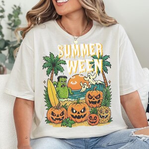 May include: Cream-colored t-shirt with a graphic that says "Summerween". The graphic features a beach scene with palm trees, pumpkins, a monster, ghosts, a surfboard, and a pineapple.