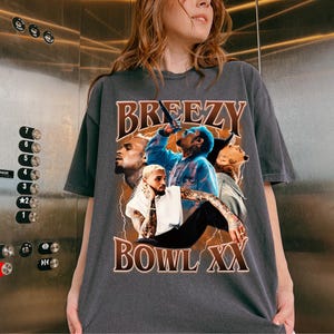 Puede incluir: Camiseta gris oscuro con un dise&ntilde;o gr&aacute;fico. La palabra "BREEZY" en letras marrones est&aacute; sobre un collage de im&aacute;genes. Las im&aacute;genes incluyen una persona con una camisa azul y otros retratos. El texto "BOWL XX" est&aacute; debajo.