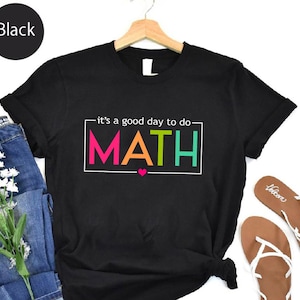 May include: Black t-shirt with the text "it's a good day to do MATH" in a colorful block design. The shirt is displayed with blue jeans, white flowers, and brown sandals.