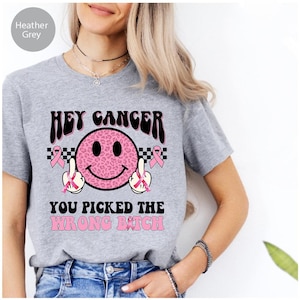 Puede incluir: Camiseta gris jaspeado con un diseño gráfico. El diseño presenta el texto "HEY CANCER" sobre una carita sonriente rosa con estampado de leopardo con los dedos medios levantados. El texto "YOU PICKED THE WRONG BITCH" está debajo.