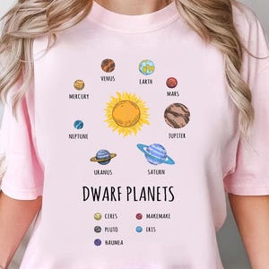 Puede incluir: Una camiseta rosa claro con una colorida ilustración del sistema solar. El diseño incluye el sol, los planetas y los planetas enanos, con el texto "DWARF PLANETS".