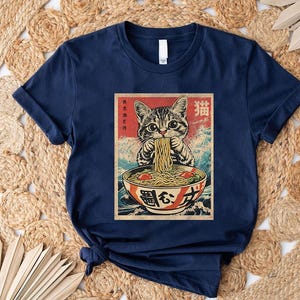 A navy blue t-shirt with a graphic design depicting a cat eating noodles from a bowl.
