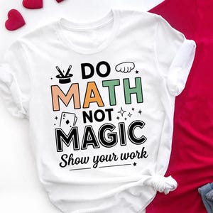 Może przedstawiać: Biała koszulka z napisem "DO MATH NOT MAGIC Show your work" w kolorze czarnym, zielonym i pomarańczowym. Projekt zawiera kapelusz magika, karty do gry i gwiazdy. Koszulka jest złożona na dole.