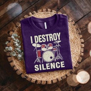 Pode incluir: Uma camiseta roxa com o texto "I DESTROY SILENCE" e um desenho de bateria em branco. A bateria inclui tambores vermelhos e ferragens prateadas. A camiseta é exibida em um sousplat circular tecido.