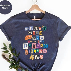 May include: Heather navy t-shirt with the text "What's More Punk Than The Public Library??" in a colorful, cut-out style. The text is arranged in a vertical stack, with each word in a different font and color. The shirt is hanging on a wooden hanger.
