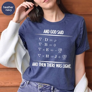 May include: Heather navy t-shirt with the text "AND GOD SAID" and mathematical equations, followed by "AND THEN THERE WAS LIGHT." The shirt is a crew neck style.