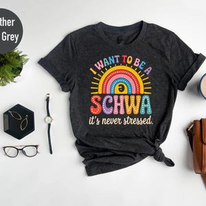 May include: Heather dark grey t-shirt with the text "I want to be a Schwa, it's never stressed." The design features a colorful rainbow and sun graphic. The shirt is a casual wear item.