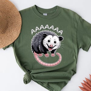 May include: Olive green t-shirt featuring a cartoon opossum with the word "AAAAAAA" above it. The opossum has black and white fur, a pink tail, and is smiling with its mouth open. The shirt is folded.