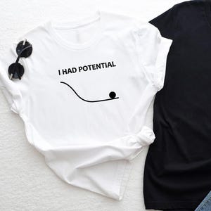 Könnte beinhalten: Weißes T-Shirt mit dem Text "I HAD POTENTIAL" über einer schwarzen Linie und einem schwarzen Kreis. Eine Sonnenbrille liegt auf der Schulter des Shirts. Ein schwarzes T-Shirt befindet sich neben dem weißen Shirt.