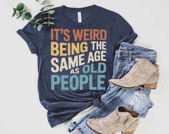Könnte beinhalten: Marineblaues T-Shirt mit dem Text "IT'S WEIRD BEING THE SAME AGE AS OLD PEOPLE" in Orange, Gelb, Weiß und Türkis. Das Shirt wird mit einer blauen Jeans und beigen Stiefeletten kombiniert.