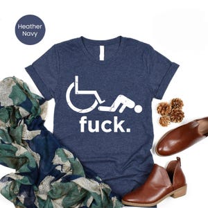 Puede incluir: Camiseta azul marino con un gráfico blanco de una persona en silla de ruedas en una posición comprometida y la palabra "fuck". La camiseta se muestra con zapatos marrones, piñas y una bufanda estampada.