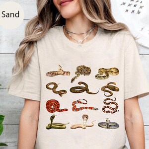 May include: A sand-colored t-shirt featuring a graphic print of twelve different snakes arranged in a grid. The snakes are depicted in various colors including brown, red, black, and yellow, showcasing diverse patterns and coiled positions.