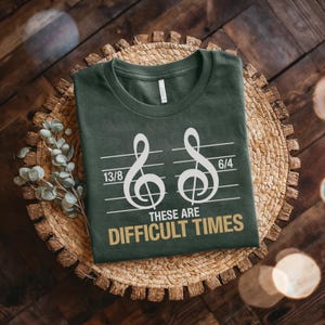 Peut inclure: T-shirt vert foncé avec deux clés de sol blanches et les chiffres 13/8 et 6/4. Le texte doré dit "THESE ARE DIFFICULT TIMES". Le t-shirt est présenté sur un panier en osier, parfait pour les mélomanes.