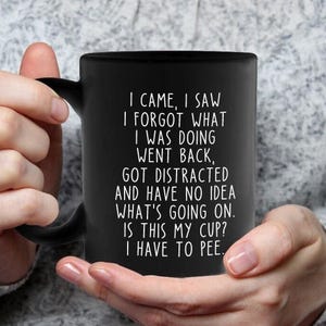 May include: Black ceramic coffee mug with a curved handle. The mug features white text that reads: "I CAME, I SAW, I FORGOT WHAT I WAS DOING, WENT BACK, GOT DISTRACTED AND HAVE NO IDEA WHAT'S GOING ON. IS THIS MY CUP? I HAVE TO PEE."