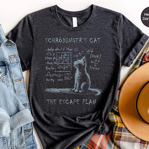 Könnte beinhalten: Dunkelgraues meliertes T-Shirt mit einer Zeichnung einer Katze und mathematischen Gleichungen. Der Text lautet "Schrödinger's Cat" und "The Escape Plan". Das T-Shirt hat einen Rundhalsausschnitt und hochgekrempelte Ärmel.