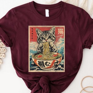 A burgundy t-shirt with a graphic design depicting a cat eating noodles from a bowl.