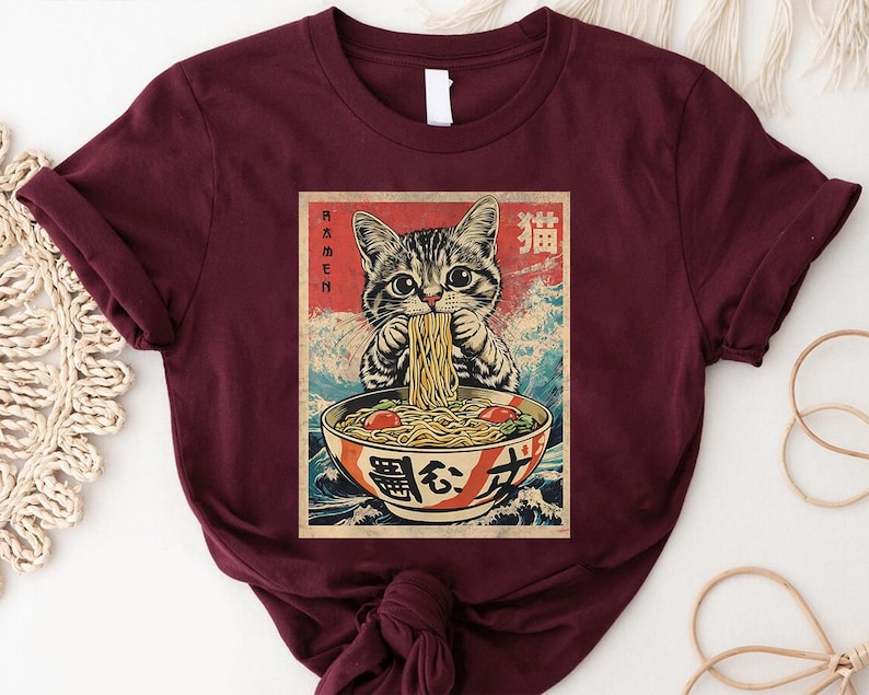 A burgundy t-shirt with a graphic design depicting a cat eating noodles from a bowl.