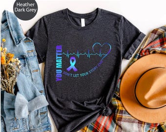 May include: Heather dark grey t-shirt with a graphic design. The design includes a ribbon, a heartbeat line, and the words "YOU MATTER" and "DON'T LET YOUR STORY END" in a gradient of purple and blue.