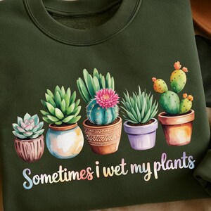 May include: Dark green sweatshirt featuring a watercolor illustration of potted succulents and cacti. The text "Sometimes I wet my plants" is written in a colorful, whimsical font. The design is centered on the front of the garment.