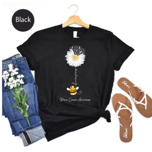 May include: Black t-shirt featuring a daisy with a yellow center and white petals, with the words "bee strong" and a cartoon bee. The shirt also has the words "Brain Cancer Awareness". The shirt is paired with blue jeans and brown sandals.