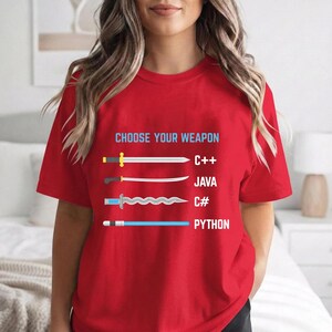A woman wearing a red t-shirt with a graphic design that says "Choose your weapon" and lists four programming languages: C++, Java, C#, and Python.