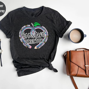 May include: Heather dark grey t-shirt with a colorful graphic. The graphic features a heart-shaped apple with the words "Assistant Director" and surrounding words like "Reliable", "Loving", "Fun", "Caring", and "Dedicated".