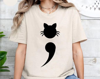 May include: Sand-colored t-shirt featuring a black cat head above a semicolon. The cat's face has pointed ears and whiskers. The shirt is made of a soft material and is suitable for casual wear.