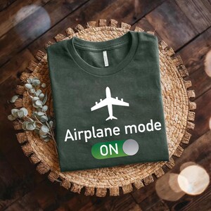 a green t-shirt with a white airplane graphic and the text "Airplane mode ON" printed on it, placed on a woven basket or plate.