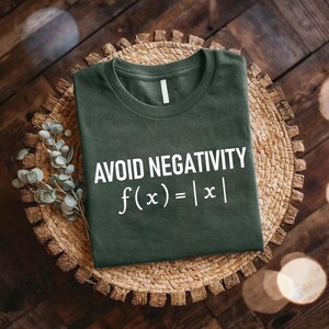 May include: Dark green t-shirt with the text "AVOID NEGATIVITY f(x) = |x|" in white. The shirt is displayed on a woven, circular placemat with a wooden background and decorative leaves.