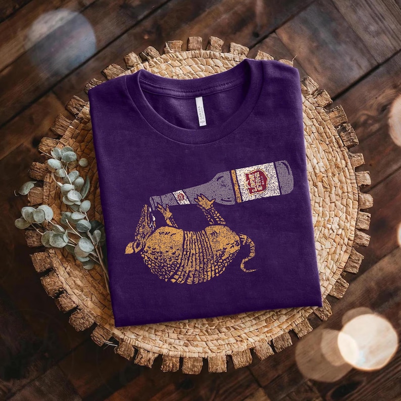 a purple t-shirt with a graphic design of a cat and a bottle of beer, placed on a woven basket or plate.