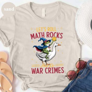May include: Sand-colored t-shirt with a whimsical graphic. The design features a frog in a wizard hat and cape riding a goose, holding a yellow die. Text reads "LET'S ROLL MATH ROCKS AND DO WAR CRIMES" in red and yellow.