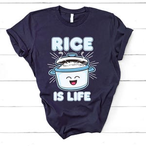 May include: A navy blue t-shirt featuring the phrase "RICE IS LIFE" and a cartoon rice cooker. The rice cooker is white and blue, with steam rising from the lid. The text is in a light blue font.
