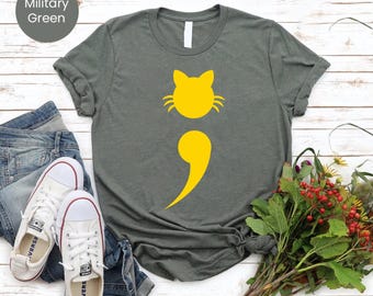 May include: Military green t-shirt with a yellow cat design. The cat design is a combination of a cat head and a semicolon. The shirt is paired with blue jeans and white sneakers.