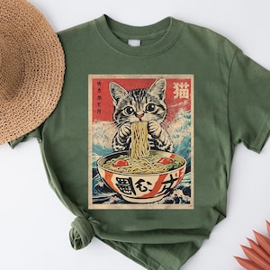 A green t-shirt with a graphic design depicting a cat eating noodles from a bowl, surrounded by Japanese text and imagery.