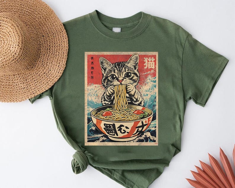A green t-shirt with a graphic design depicting a cat eating noodles from a bowl, surrounded by Japanese text and imagery.