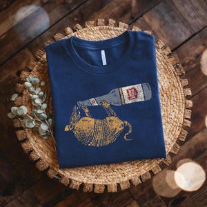 A navy blue t-shirt with a graphic design of a cat holding a beer bottle is displayed on a woven basket.