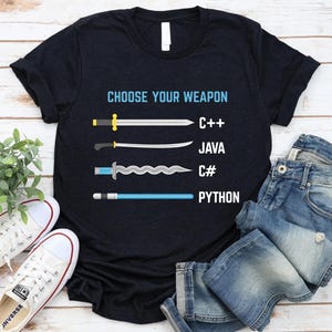 a black t-shirt with a graphic design that says "Choose Your Weapon" and lists four programming languages: C++, Java, C#, and Python, each represented by a different type of sword. The t-shirt is laid out on a wooden surface, with a pair of jeans and white sneakers visible in the background.