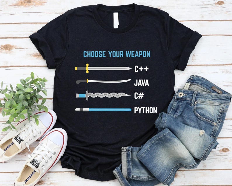 a black t-shirt with a graphic design that says "Choose Your Weapon" and lists four programming languages: C++, Java, C#, and Python, each represented by a different type of sword. The t-shirt is laid out on a wooden surface, with a pair of jeans and white sneakers visible in the background.