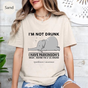 May include: Sand-colored t-shirt with the text "I'M NOT DRUNK" above a gray cartoon elephant. Below the elephant, the text reads "I HAVE PARKINSON'S OKAY... MAYBE I'M A 'LIL DRUNK" with the hashtag #parkinson's-awareness.