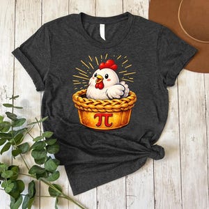 May include: Dark gray t-shirt featuring a cartoon chicken in a pie crust with the pi symbol. The chicken has a red comb and beak, and the pie crust is golden brown. The design includes yellow rays emanating from behind the chicken.