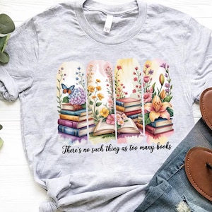 May include: A light grey t-shirt with a vibrant graphic of books and flowers. The design includes a butterfly and the words "There's no such thing as too many books". The shirt is paired with denim jeans.