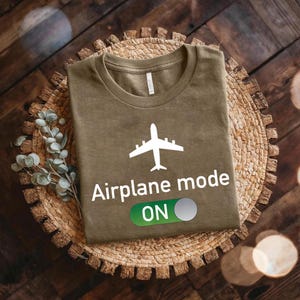 a brown t-shirt with a white airplane graphic and the text "Airplane mode ON" printed on it, placed on a woven basket or plate.