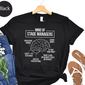May include: Black t-shirt featuring a white diagram of a stage manager's mind, with text including 'backstage drama' and 'tech week'. The shirt is displayed with blue jeans and sandals.