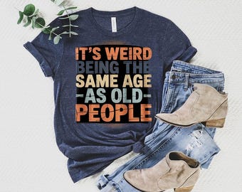 Könnte beinhalten: Marineblaues T-Shirt mit dem Text "IT'S WEIRD BEING THE SAME AGE AS OLD PEOPLE" in einem farbenfrohen Retro-Design. Dazu blaue Jeans und beige Stiefeletten.