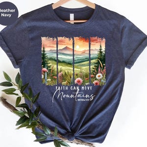 May include: Heather navy t-shirt featuring a watercolor landscape with mountains, flowers, and a sunset. The text "Faith Can Move Mountains" is displayed below the image, with the Bible verse Matthew 17:20. The shirt is hanging on a wooden hanger.